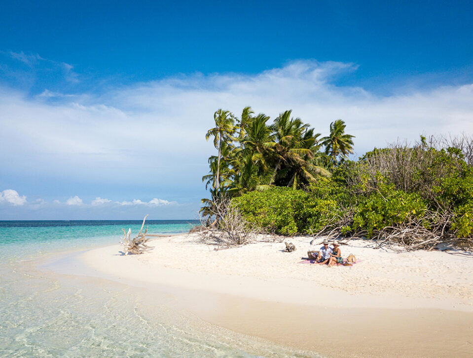 Seychelles: A couple relaxes on a small sandy beach with clear turquoise water, surrounded by lush green palm trees and bushes under a bright blue sky with scattered clouds.