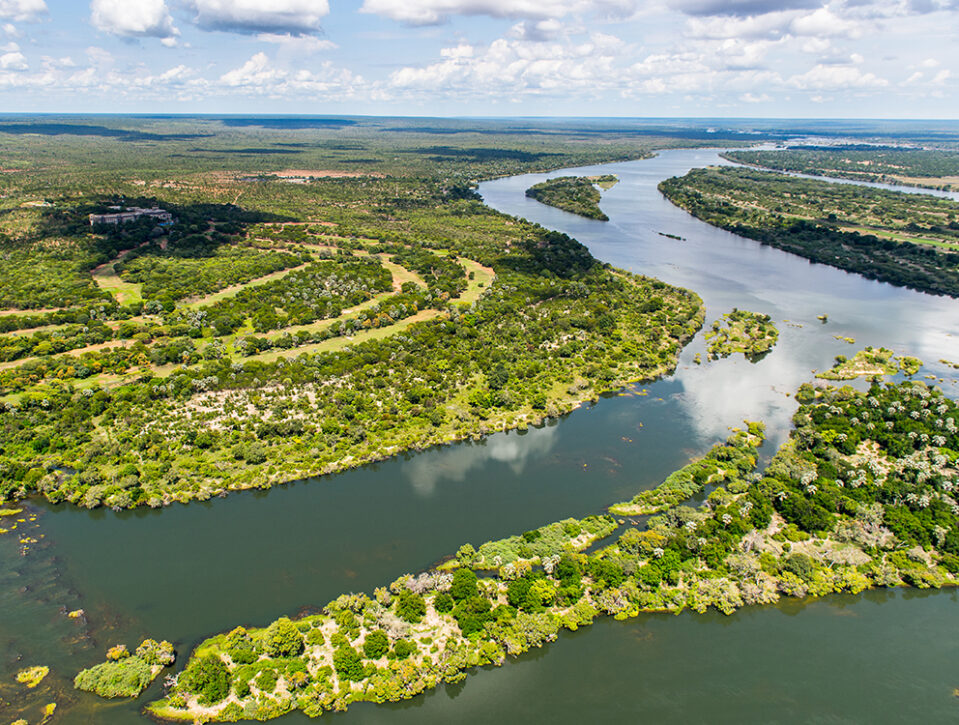Zambia: Aerial view of a wide, winding river bordered by lush green vegetation and islands, under a partly cloudy sky with expansive forest in the background.