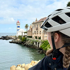 Portugal: A person wearing a bike helmet looks toward a lighthouse and historic buildings by the sea.
