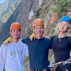 Peru: Three people wearing helmets and safety gear stand together on a rocky mountain trail, smiling at the camera with cliffs and greenery in the background.