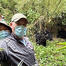 Rwanda: Two people wearing masks and outdoor gear take a selfie in a lush, green forest with a group of gorillas sitting on the ground in the background.