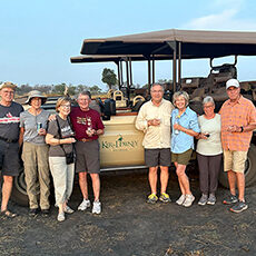 A family stands in front of a safari vehicle, smiling and holding drinks, with dry grass and trees in the background.