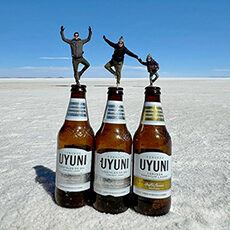 Three people pose in yoga stances, appearing to stand on top of three Uyuni beer bottles, with a flat, expansive salt plain in the background.
