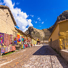 Peru: Cobblestone street lined with colorful textiles and crafts displayed on tables outside rustic yellow and stone buildings, with mountains and a blue sky in the background.