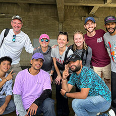 A group of nine people, including Kathy Sharpe, pose together indoors, smiling at the camera. Some are kneeling in front, while others stand behind them. Casual clothing and hats are worn by several individuals.