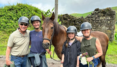 The rise of grandparents traveling with grandchildren: Four people wearing helmets stand next to a brown horse outdoors, smiling at the camera. A stone wall and greenery are visible in the background.