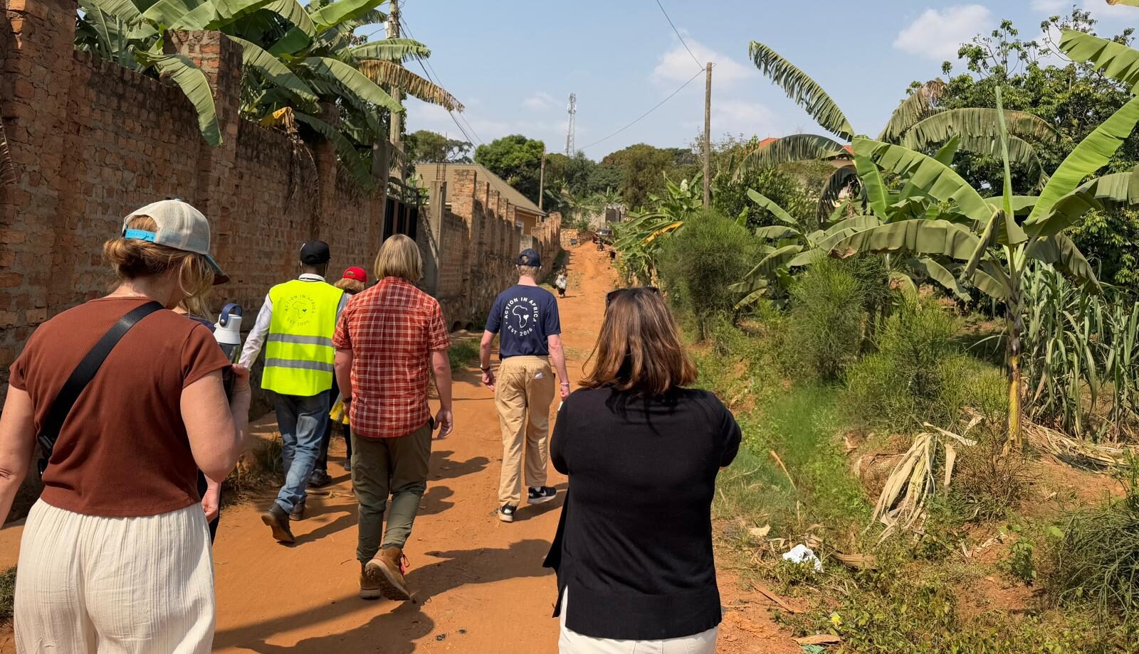 a group on a donor trip walk down a dirt road with a brick wall on one side and green foliage on the other in Rwanda