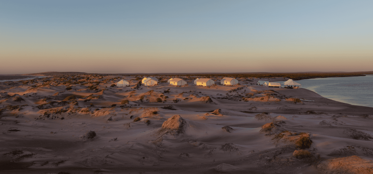tents for a group donor trip itinerary amidst an isolated sandy beach beside the ocean at sunset.
